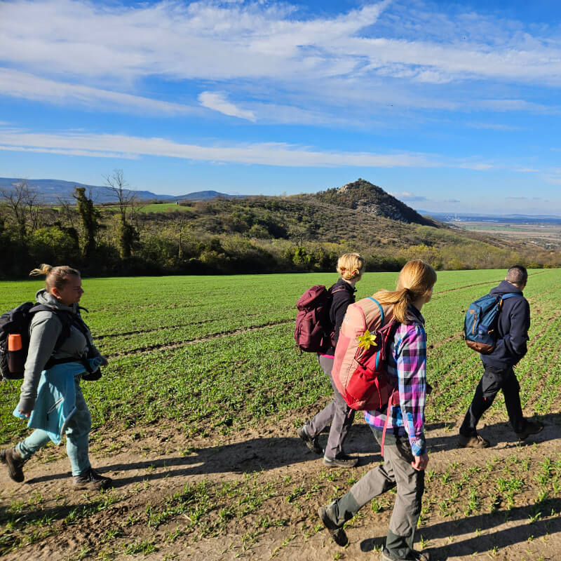 Turisták haladnak egy szántóföld szélén, háttérben hegyek, dombok.